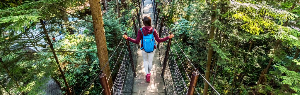 Girl dress in maroon jacket, white leggings, and pink shoes walking across a bridge on a hiking trail in the woods, vancouver victoria performance tours
